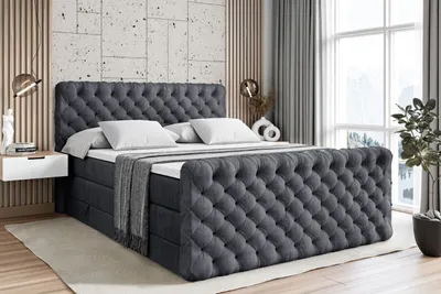 A dark gray tufted boxspring bed with white pillows and a gray throw blanket is shown in a bedroom setting.