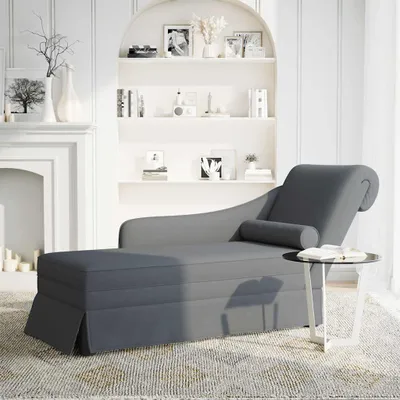 A dark gray chaise lounge with a bolster pillow is placed in a living room setting next to a fireplace and a bookshelf.