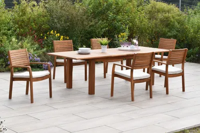 A wooden dining set with chairs and a table is placed on a patio surrounded by greenery.