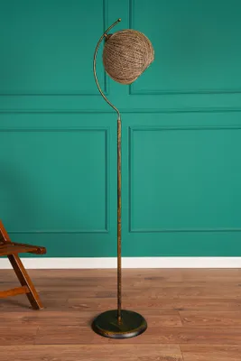 A floor lamp with a jute shade stands on a wooden floor in front of a teal wall.