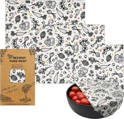A set of beeswax food wraps with a vegetable pattern, displayed with packaging and a bowl of tomatoes.