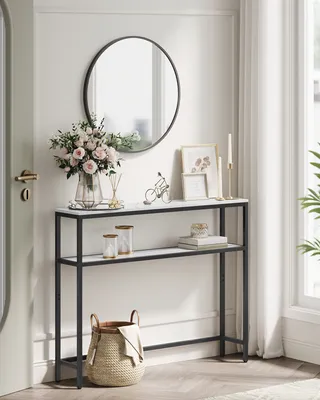 A narrow console table with two shelves, decorated with flowers, picture frames, and a basket, placed in a hallway.