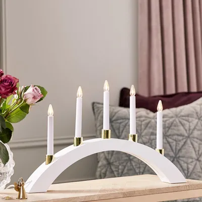 A white arched LED candle bridge with five warm white LED lights sits on a wooden surface, with a vase of roses to the left and a patterned cushion and draped fabric in the background.