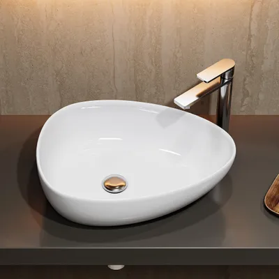 A white oval vessel sink with a chrome faucet and a gold drain sits on a dark gray countertop against a textured tan wall.