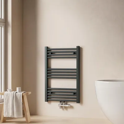 A dark grey towel radiator mounted on a light beige wall, next to a window and a wooden stool with a folded towel.