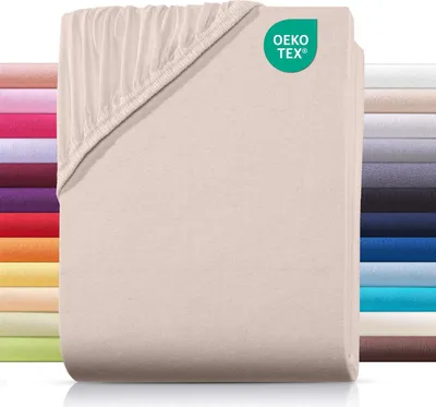 A stack of folded beige fitted sheets with a corner showing the elasticized edge. A row of other colored sheets is visible on the left, and a stack of different colored sheets is visible on the right.