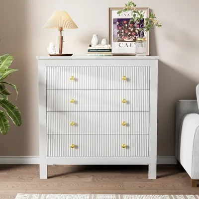 A white dresser with gold knobs is shown in a living room setting, next to a sofa and a plant.