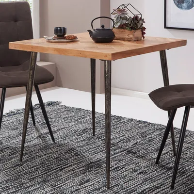 A square wooden dining table with metal legs, set on a patterned rug, with two chairs partially visible.