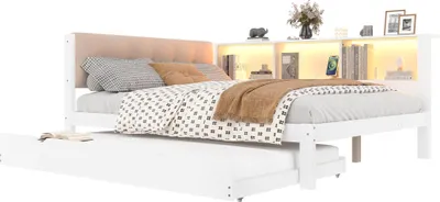 A white wooden bed with a beige upholstered headboard and a pull-out trundle bed underneath. The headboard has integrated shelving with LED lighting, displaying decorative items and a smartphone.