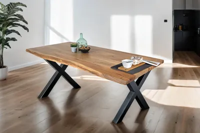 A dining table with a live edge and black metal X-shaped legs, set in a modern dining room with wooden floors and a large plant.