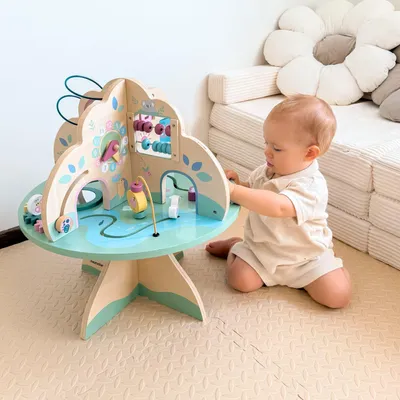 A baby is playing with a wooden activity table in a room with a large flower-shaped cushion in the background.