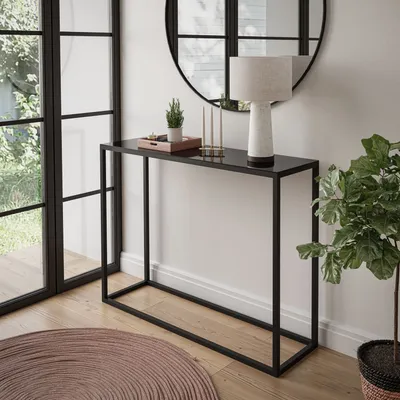 A modern console table with a black glass top and black metal frame, decorated with a small plant, candles, and a lamp. It is placed in a room with large windows and a round rug.