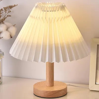 A close-up of a white pleated table lamp with a wooden base and stem, placed on a white surface.
