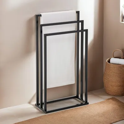 A black metal towel rack with three bars, holding white towels, stands against a light-colored wall. A woven basket with towels is visible to the right.