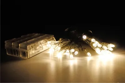 A string of warm white LED lights with a battery pack, displayed on a reflective surface against a dark background.