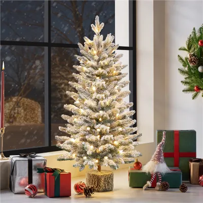 A small, flocked Christmas tree with lights is displayed on a surface with gifts and decorations, with a snowy window in the background.
