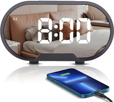 A digital alarm clock with a mirrored surface displaying the time, placed in front of a bedroom scene. A smartphone is shown charging wirelessly in the foreground.
