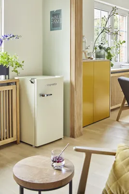 A small cream-colored refrigerator is placed in a room next to a wooden radiator and a yellow cabinet. A small round table with a glass of dessert is in the foreground.