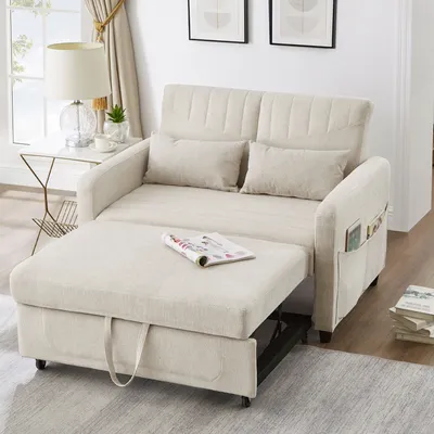 A beige sofa bed is shown in a living room setting, with a side table and lamp to the left, and a stack of books to the right.