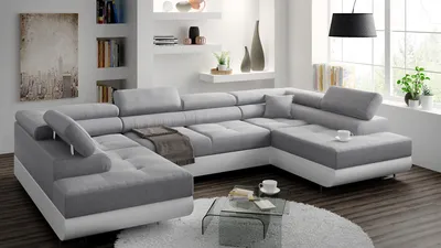 A large, sectional sofa in a modern living room setting. The sofa is gray with white accents and sits on a wooden floor with a white rug. There are decorative items on shelves in the background.
