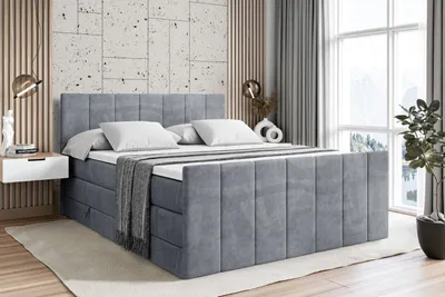 A bedroom scene featuring a gray upholstered bed with white pillows and a gray throw blanket. The bed is placed on a rug in front of a textured wall and large windows.