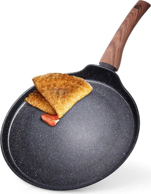 A close-up, slightly angled view of a black crepe pan with a granite coating and a wooden handle. Two folded crepes and a slice of strawberry are on the pan.