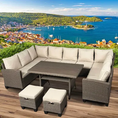 A polyrattan garden seating group with a table and ottomans is set up on a wooden deck, with a scenic coastal town and bay in the background.