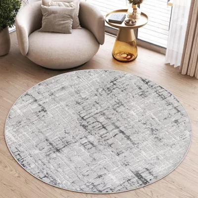 A round rug with a modern abstract pattern is placed on a wooden floor in a living room setting, next to a comfortable armchair and a side table.