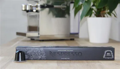 A Nespresso coffee capsule and its packaging are displayed on a wooden surface in front of a coffee machine and a potted plant.