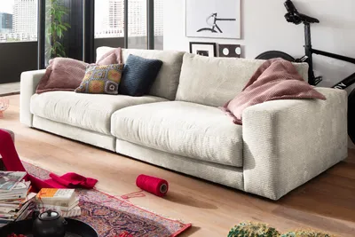 A modern living room with a large, cream-colored corduroy sofa as the main focus. Several decorative pillows are arranged on the sofa, and a pink throw blanket is draped over one armrest. In the foreground, there's a rug with a patterned design, a stack of