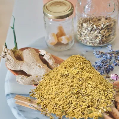 A pile of yellow powder, a ginger root, and dried lavender are arranged on a marble surface with two glass jars in the background.