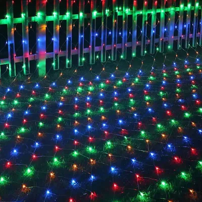 A net of colorful lights is spread across grass in front of a wooden fence.