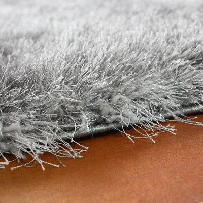 A close-up view of the edge of a shaggy, high-pile rug with silver or gray fibers resting on a brown surface.