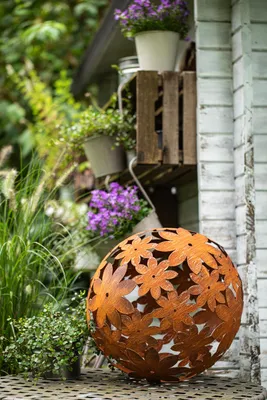 A rusty metal sphere with a floral cut-out pattern sits on a patterned surface in a garden setting, with plants and a wooden shelf in the background.