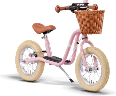 A pink balance bike with a brown seat and handlebars, and a wicker basket on the front, is shown against a white background.
