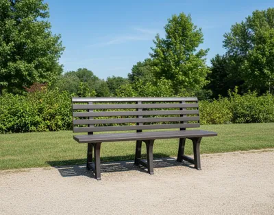 A taupe-colored, three-seater bench made of plastic sits on a gravel path in a park-like setting with trees and green bushes in the background under a clear blue sky.