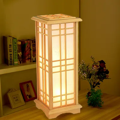A wooden floor lamp with a parchment shade is placed on a wooden surface next to a bookshelf and a vase of flowers.