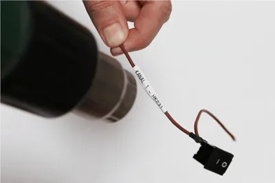 A hand holds a wire with a label and a switch attached to it, with a dark object in the background.
