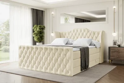 A luxurious beige velvet boxspring bed with a tufted headboard and footboard is set up in a well-lit bedroom with sheer curtains and a large mirror reflecting the ceiling.