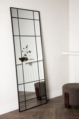 A tall, rectangular mirror with a black grid frame stands against a white wall. A small decorative plant is reflected in the mirror. A round, brown ottoman with wooden legs is placed to the right of the mirror.