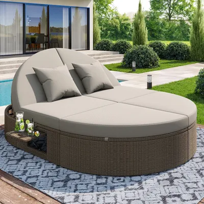 A round, rattan daybed with beige cushions and pillows is set up on a patio next to a swimming pool, surrounded by a landscaped garden.