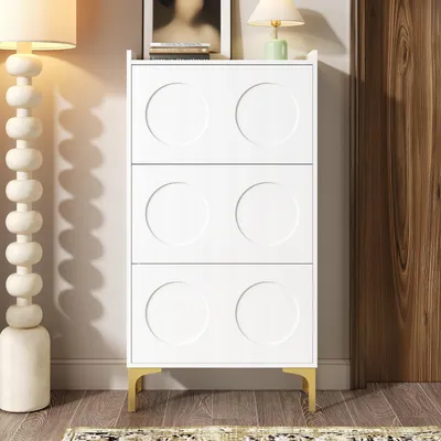 A white shoe cabinet with circular door fronts and gold legs, placed in a room with a floor lamp and a wooden door.
