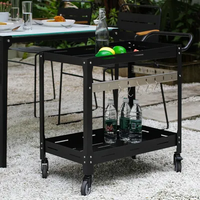 An outdoor serving cart with bottles of water and fruit on it, placed on a gravel patio next to a table.