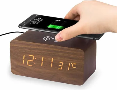 A hand places a smartphone on a wooden digital alarm clock with wireless charging capabilities.