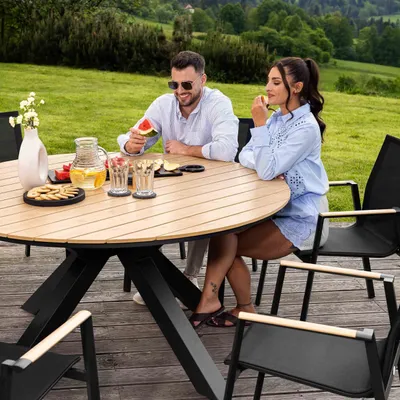 A couple enjoying a meal outdoors at a round wooden table with black chairs.