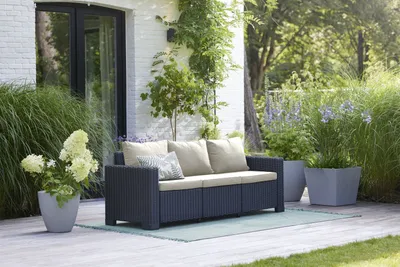 A dark grey outdoor sofa with sand-colored cushions sits on a wooden deck, surrounded by lush greenery and potted plants.