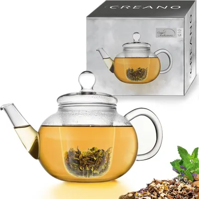 A glass teapot filled with tea, placed on a white background, with a box in the background showing the same teapot. Some loose tea leaves and mint leaves are visible in the foreground.