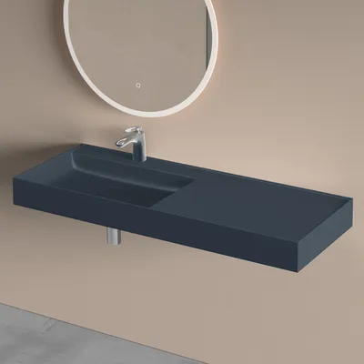 A modern, wall-mounted bathroom sink in a dark blue matte finish is shown against a light beige wall. A chrome faucet is installed in the sink, and a round, illuminated mirror is visible above it. The floor in the foreground is a light gray concrete.