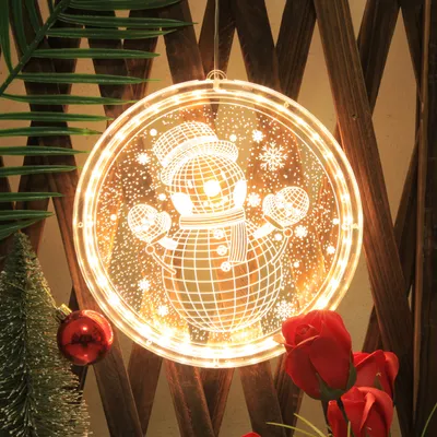A festive Christmas decoration featuring a glowing snowman light hanging on a wooden fence, with a small Christmas tree and red ornaments in the foreground.