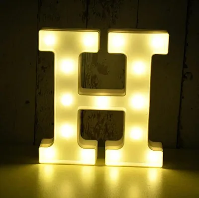 A white LED letter 'H' with illuminated bulbs is placed on a surface in front of a textured, dark background.